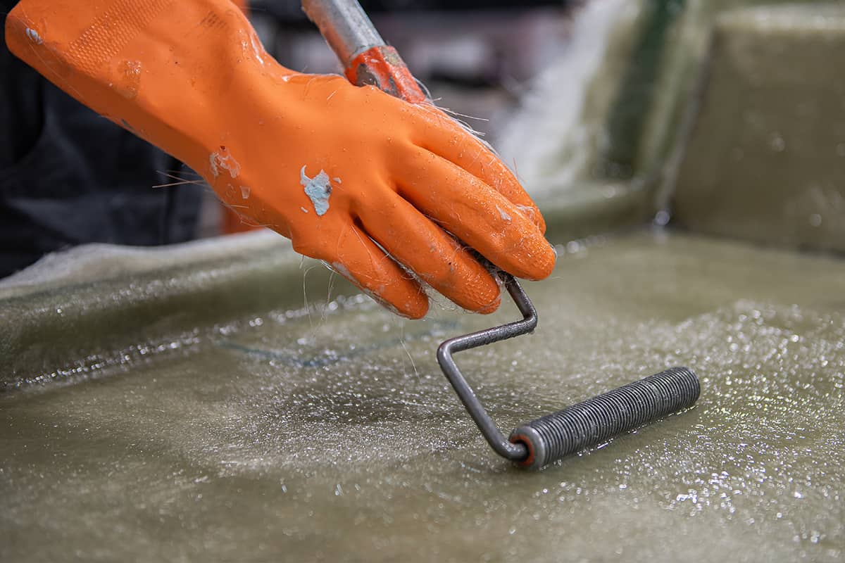 Hand of a factory worker in a glove rolling chemical solution with a tool on the fiberglass mold