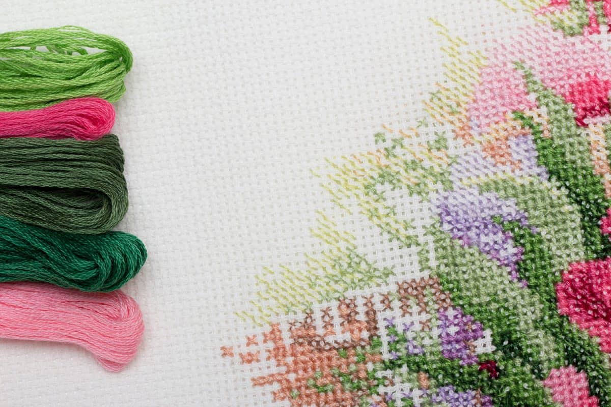 A bunch of colorful cotton embroidery threads on white aida with a fragment of embroidered flowers